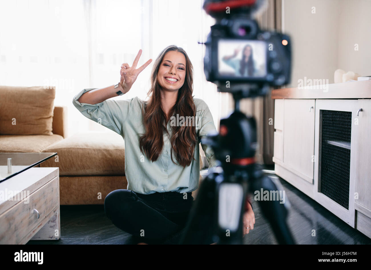 Jeune femme l'enregistrement de son blog vidéo quotidienne sur un trépied caméra installée. Souriante jeune femme montrant la victoire ou signe de paix sur l'appareil-photo. Banque D'Images