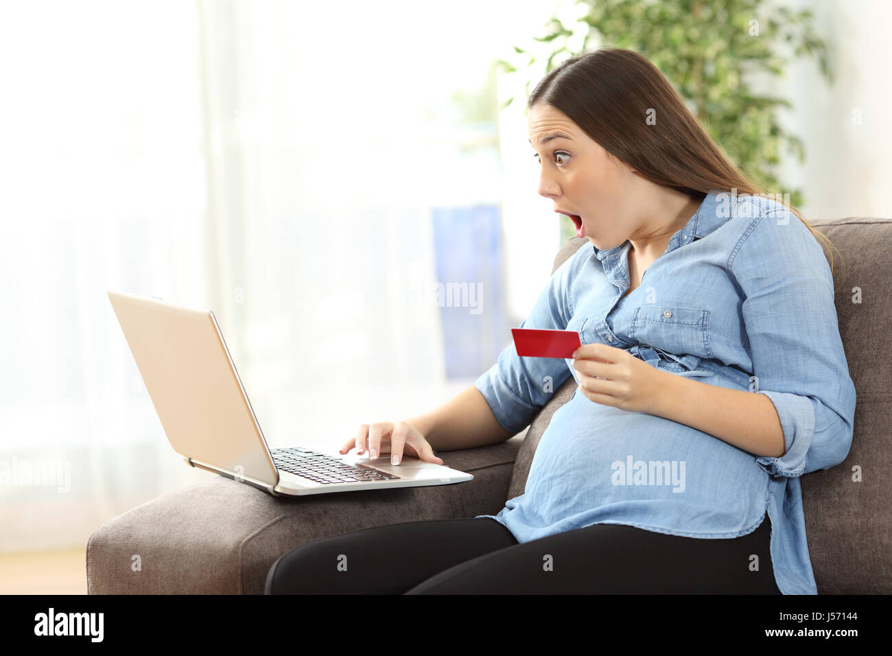 Surpris femme enceinte de vos achats en ligne par carte de crédit et d'un ordinateur portable assis sur un canapé dans le salon dans une house interior Banque D'Images
