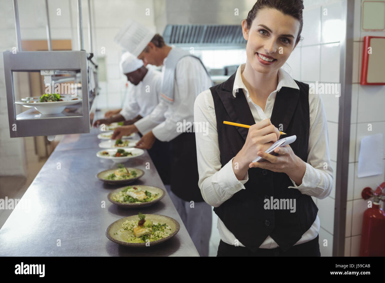 Portrait of smiling waitress avec bloc-notes de cuisine commerciale et chefs preparing food in background Banque D'Images