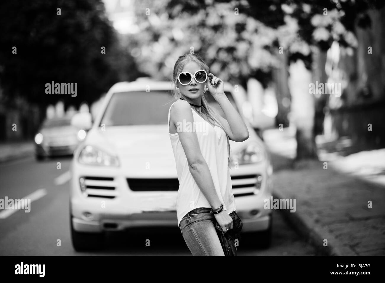 Femme blonde élégant à porter des jeans, des lunettes, collier et chemise blanche contre la voiture de luxe. Modèle urbain mode portrait. Banque D'Images