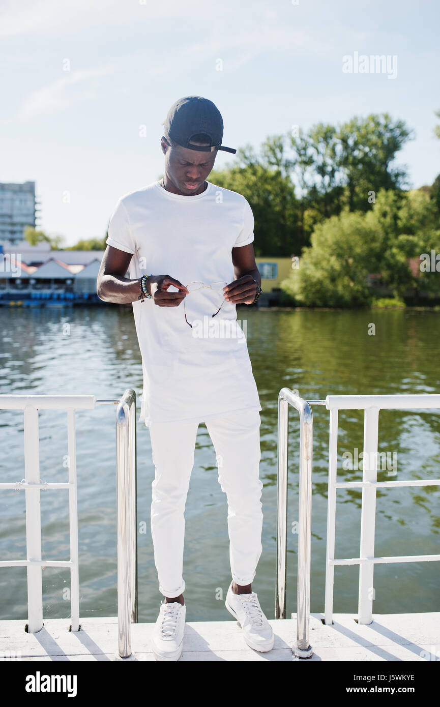 African American boy élégant, porter sur des vêtements blancs, les verres et les contre pier du lac. Mode de rue de jeunes noirs. Banque D'Images