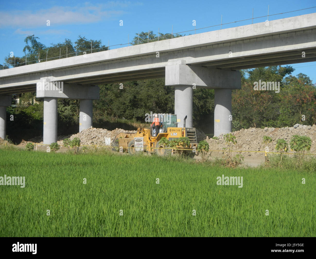 Cette image montre la phase II du projet de contournement routier artériel en cours à San Rafael, Bulacan, qui se concentre sur la transformation de l'infrastructure de regionâ€™s. L'image comprend des rizières, avec le développement routier vu en arrière-plan, illustrant le développement des systèmes agricoles et de transport. Banque D'Images