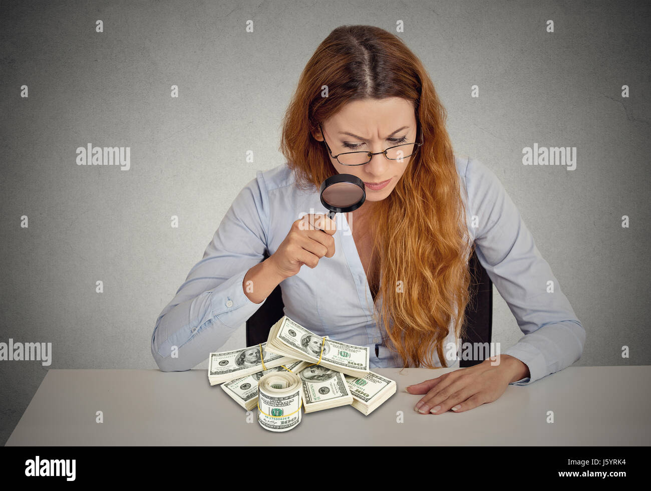 Penny Pincher. Femme d'affaires à la loupe par pile sur les billets de dollar sur fond gris table bureau isolé. Économie w Banque D'Images
