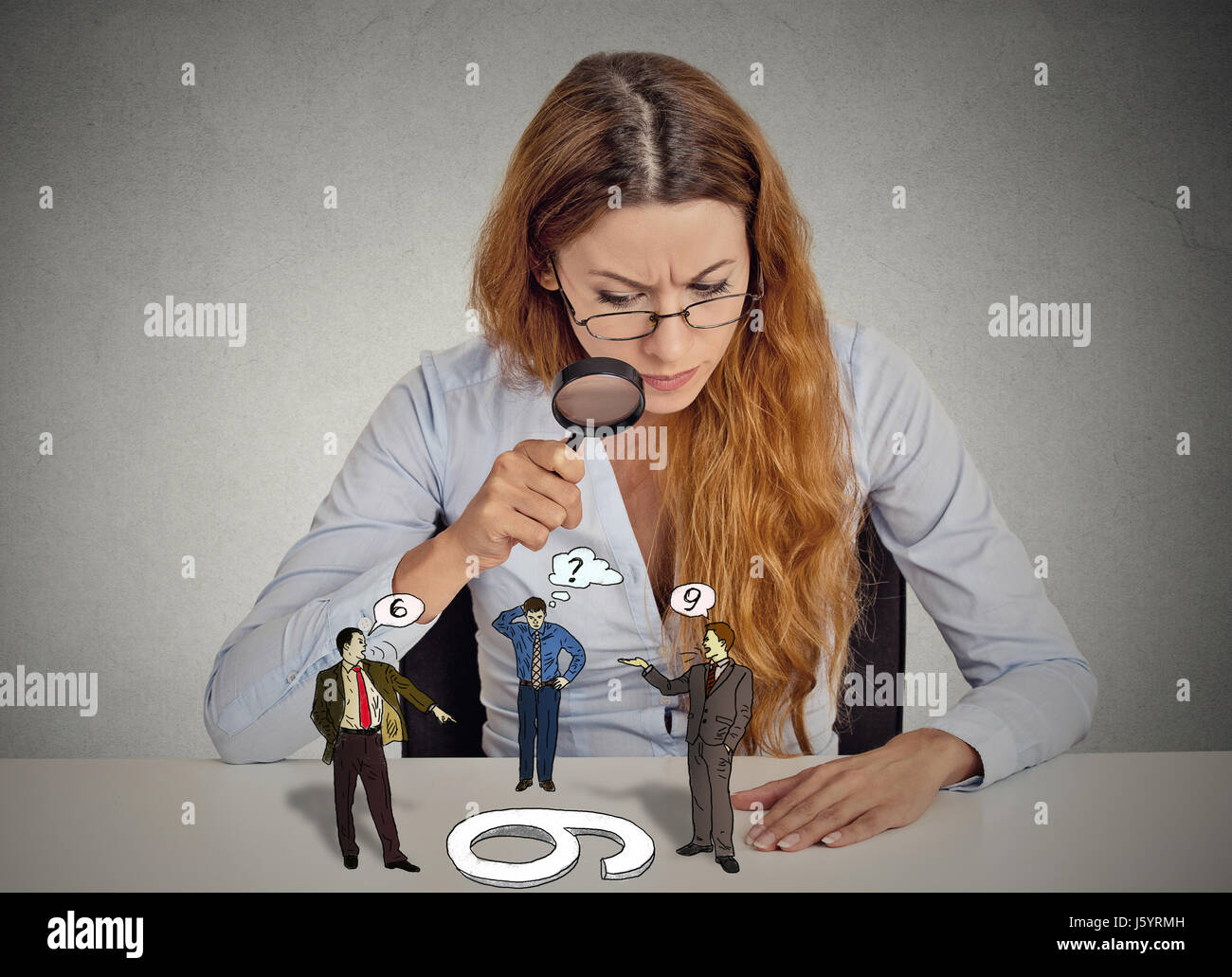 Woman with glasses sitting at desk avec scepticisme à la population à discuter par loupe bureau gris fond Mur isolé. Les droits de l Banque D'Images