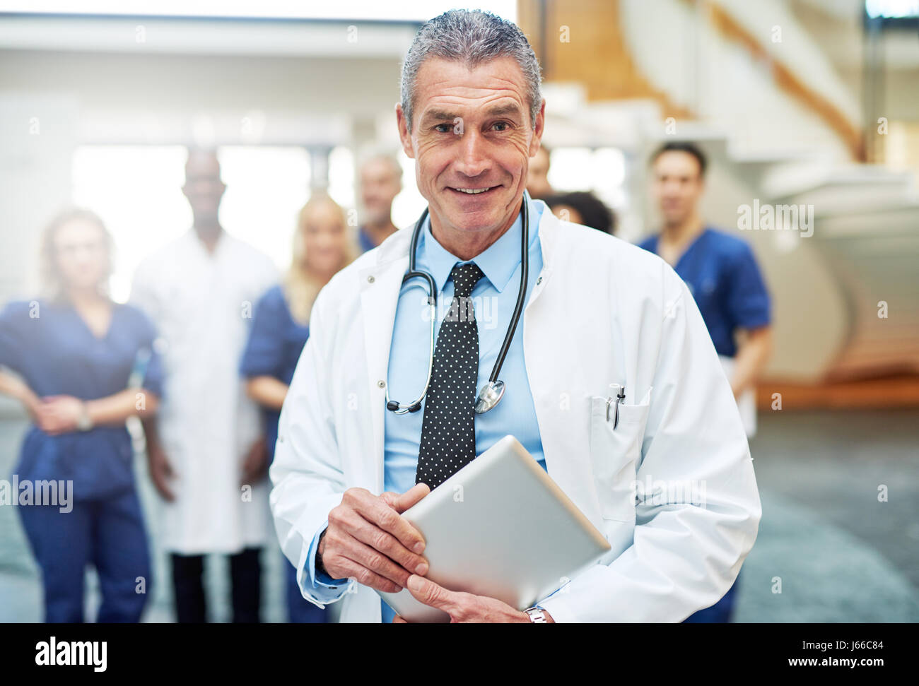 Sourire confiant friendly doctor standing avec une tablette et looking at camera dans un hôpital. Debout derrière l'équipe médicale médecin Banque D'Images