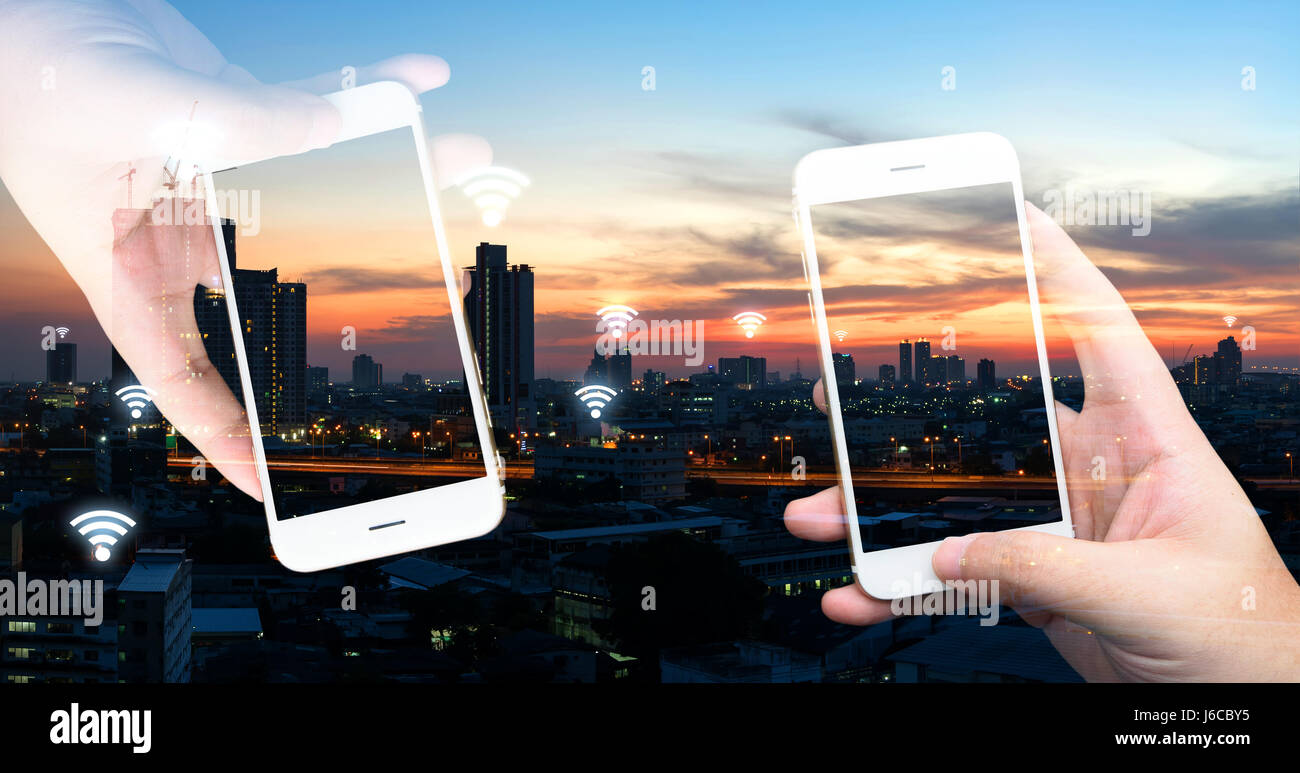 Internet des objets , iot , smart home , des ville et réseau connexion concept. Hands holding white téléphones mobiles et wifi avec ville vue du coucher de soleil. Banque D'Images