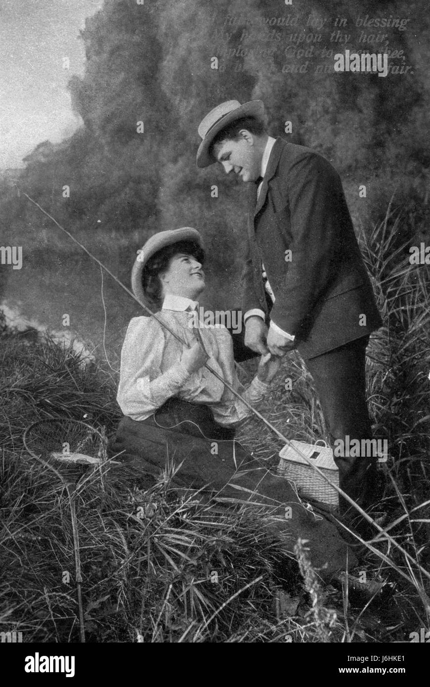 Une femme, tenant une tige de pêche en bambou (avec une ligne extrêmement lâche) et assis dans l'herbe à côté d'un étang, regarde un homme qui tient sa main. Sur l'herbe autour de la femme sont un filet de poisson manipulé avec un poisson dedans et un créel, un panier tissé que les pêcheurs ont mis leur poisson pêché dans pendant la pêche. C. 1905. Pour voir toutes mes images d'époque Alay - dans la recherche, saisissez ces 2 mots : Préstor vintage Banque D'Images