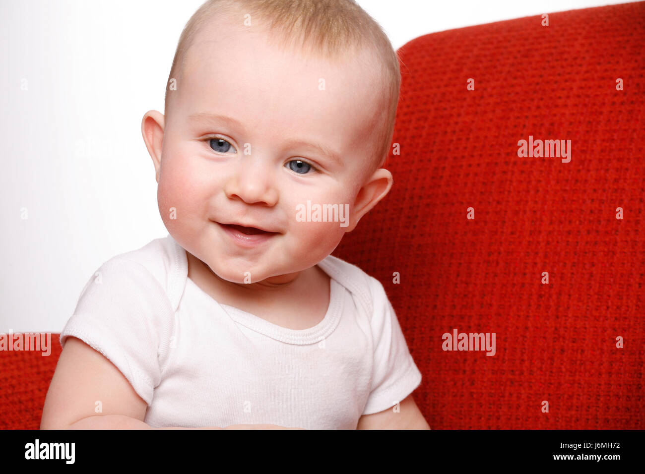 Heureux à l'âge de six mois, bébé garçon assis sur un canapé rouge, rendant les expressions faciales. Banque D'Images