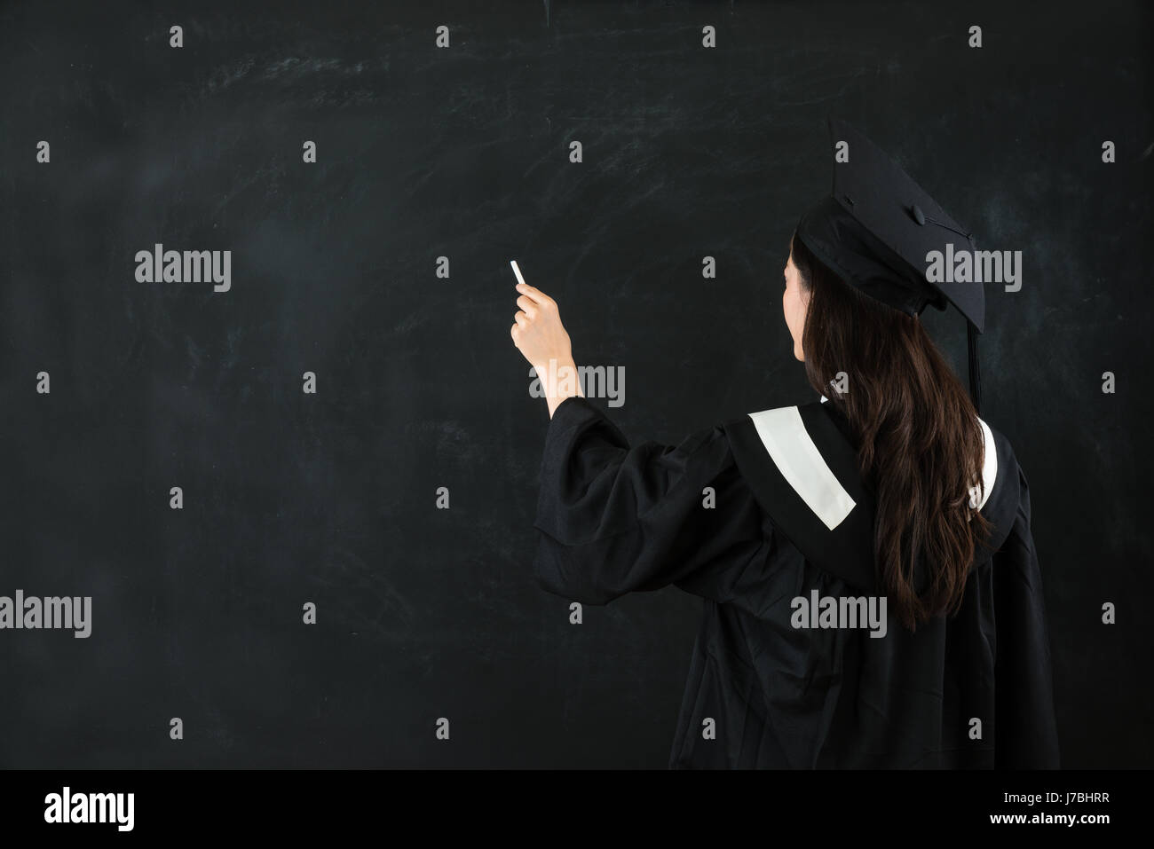 Portrait de longs cheveux en arrière à l'aide d'étudiant d'écrire du texte sur chalk board avec copyspace portant des vêtements de deuxième cycle sur fond de mur noir. Banque D'Images