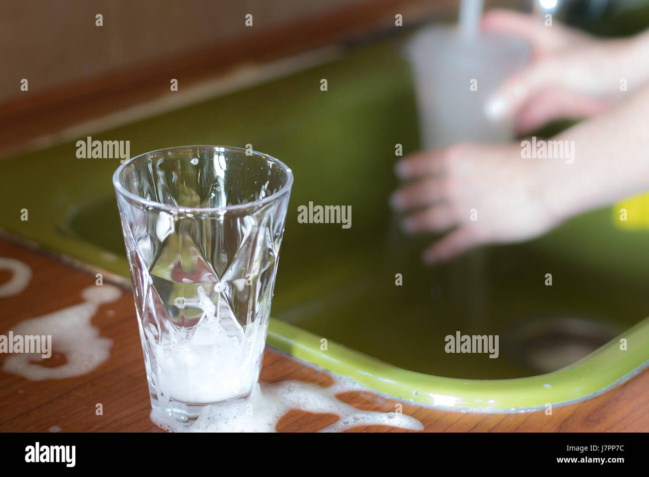 Un verres transparents avec un détergent prêt à être lavés sur un évier de cuisine avec de l'eau qui coule sur les mains floues en arrière-plan. Banque D'Images