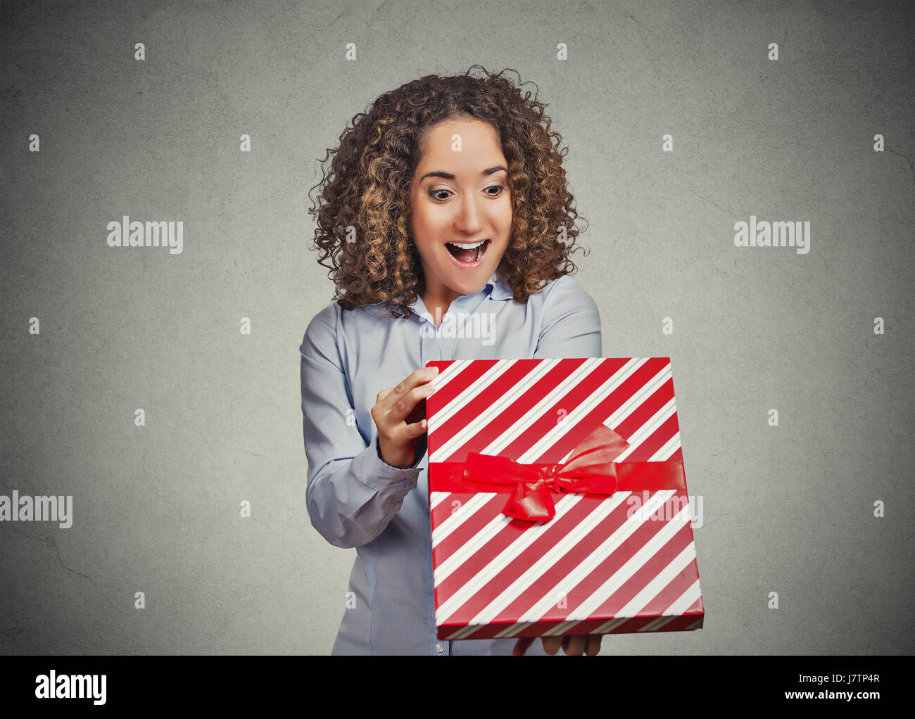 Closeup portrait heureux super excitée jeune femme ouvrant déballer le cadeau d'anniversaire rouge fort, mur gris isolé. Des émotions positives, AEC Banque D'Images