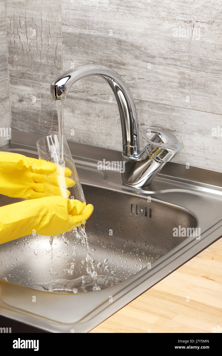 Close up de femmes dans les mains des gants en caoutchouc de protection jaune lave-verre à boire à l'eau courante contre l'évier de cuisine Banque D'Images