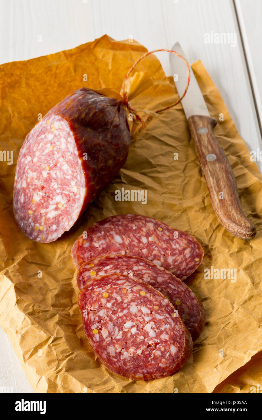 Couper le salami de boeuf sur papier d'emballage brun sur blanc table en bois avec couteau Banque D'Images