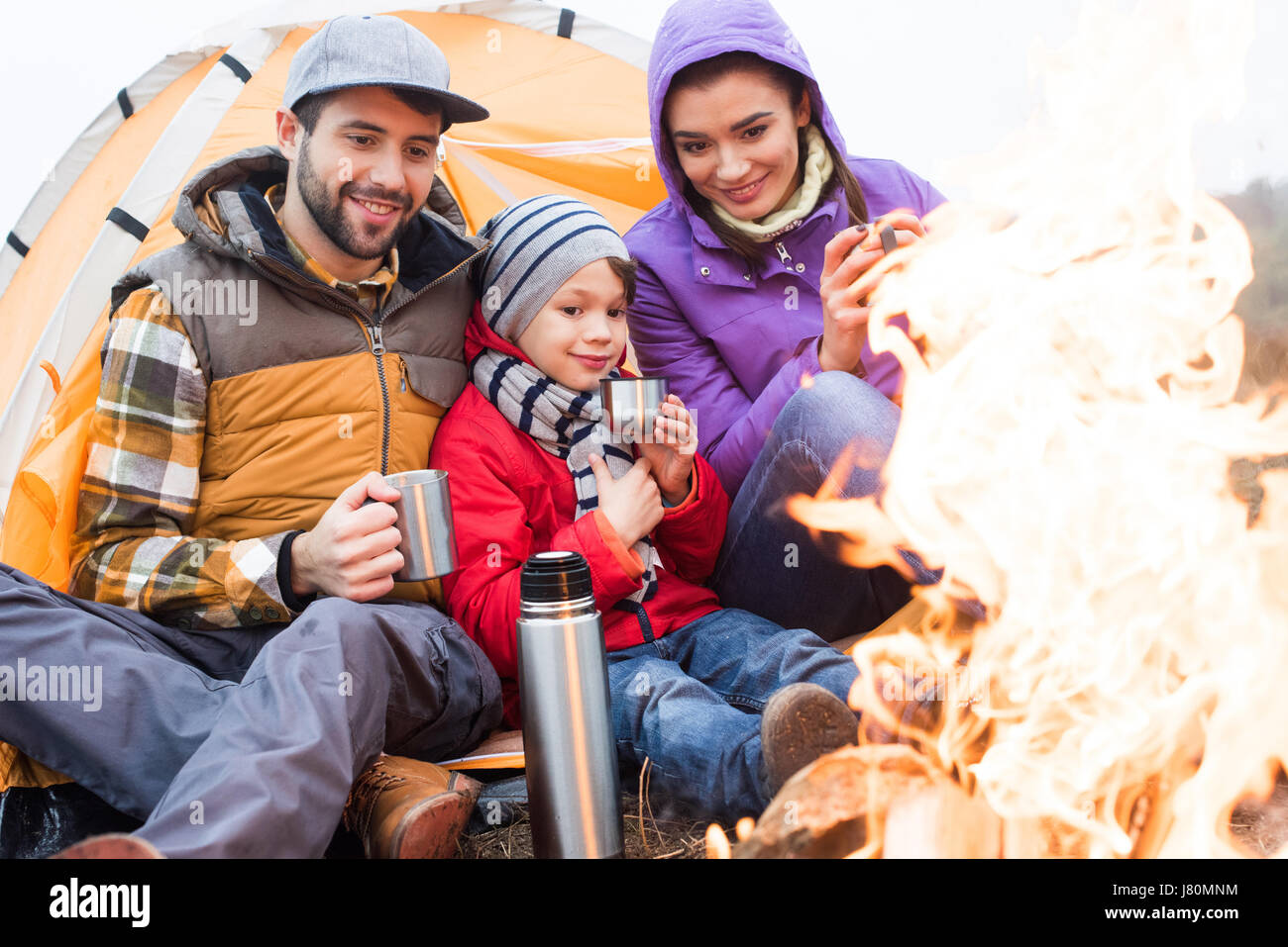 Famille d'un enfant assis près de tente et de boire le thé chaud de thermos au repos près d'un feu brûlant dans la campagne Banque D'Images
