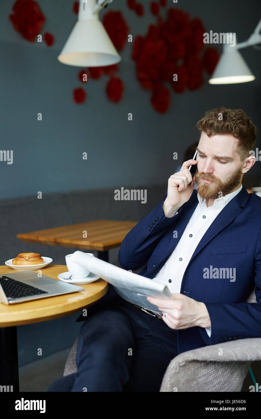 Portrait of handsome young businessman speaking par smartphone pendant la lecture de journal du matin au petit-déjeuner dans le café Banque D'Images