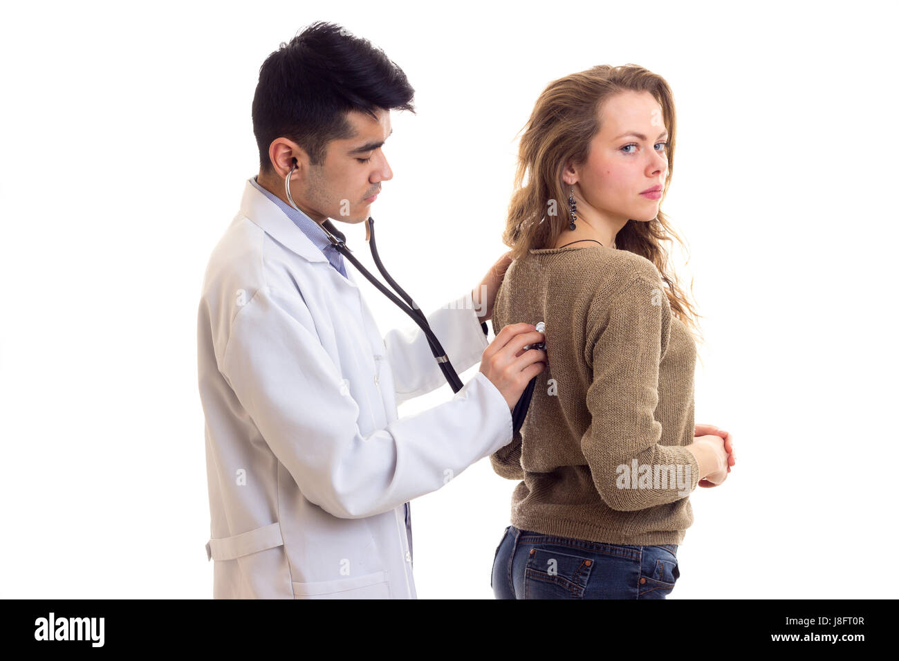 Beau jeune médecin aux cheveux noirs en robe blanche avec stéthoscope à l'écoute de l'arrière de jeune femme charmante avec de longs cheveux bruns en chandail et Banque D'Images