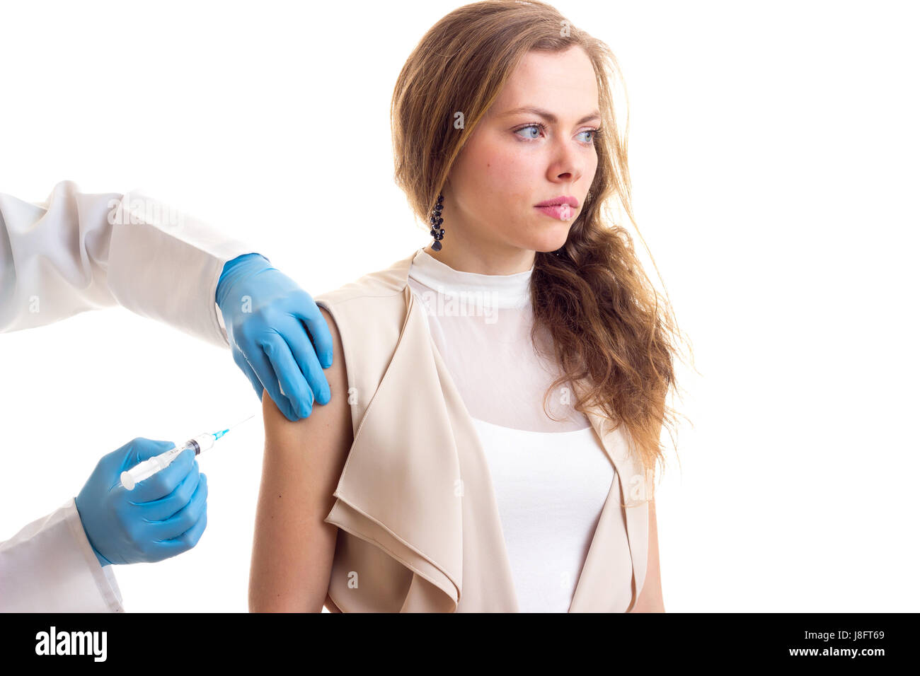 Jeune médecin en blouse blanche et gants bleu faire une injection de jeune belle femme aux longs cheveux bruns en blouse beige sur fond blanc en studio Banque D'Images