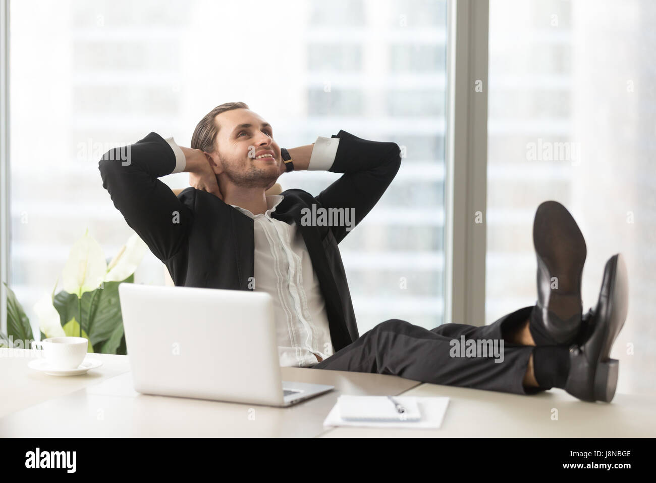 Smiling businessman looking up, assis avec les jambes sur le bureau et les mains derrière la tête. Entrepreneur de penser à des objectifs, des perspectives alléchantes, Banque D'Images
