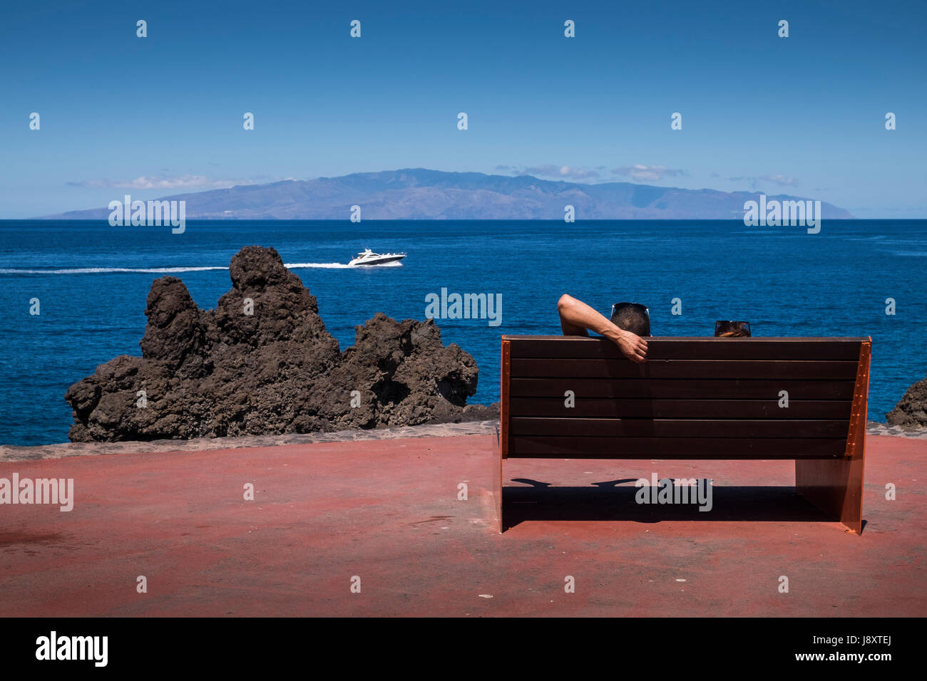 Couple relaxing on bench sur chemin côtier en regardant un bateau de pouvoir passer par sur la mer entre Tenerife et La Gomera sur une journée ensoleillée, Canaries, Banque D'Images