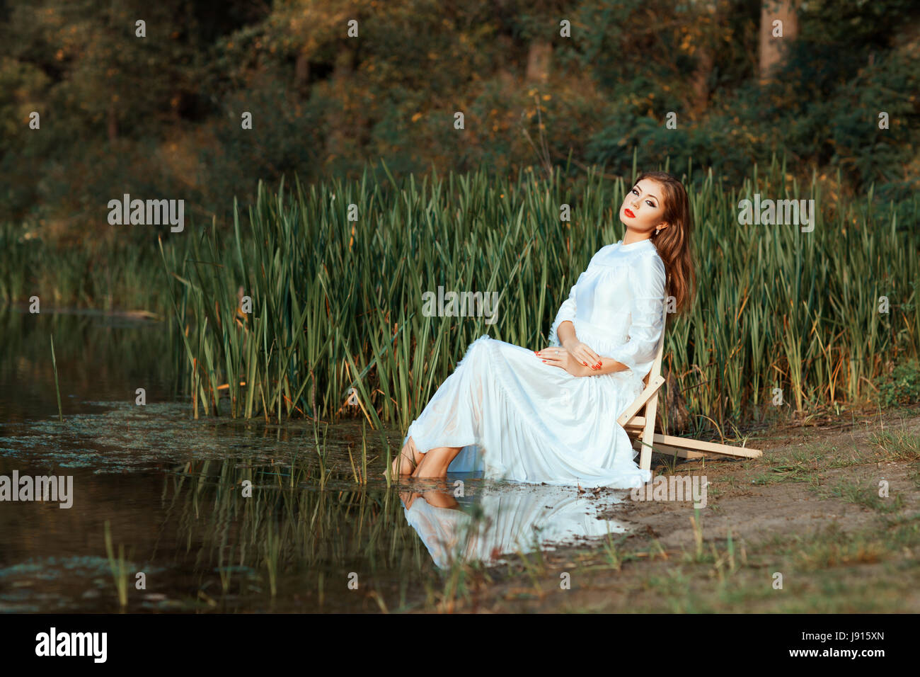 Jeune fille en robe blanche assise pieds plongeant dans le lac. Cette une journée d'été. Banque D'Images
