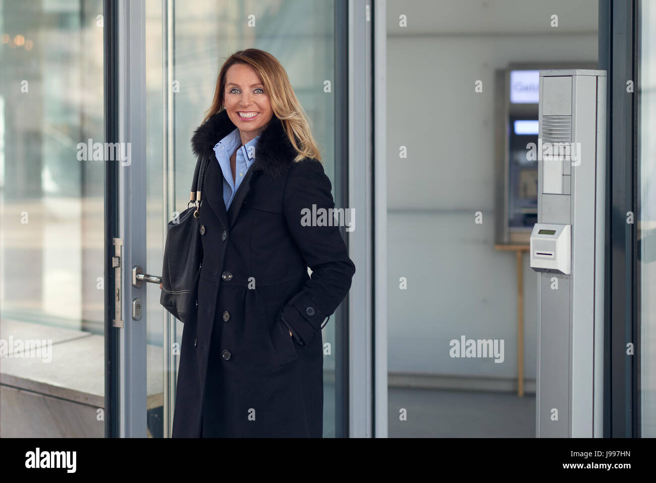 Attractive businesswoman debout dans le hall d'un immeuble commercial avec ses mains dans les poches de son pardessus smiling at the camera Banque D'Images