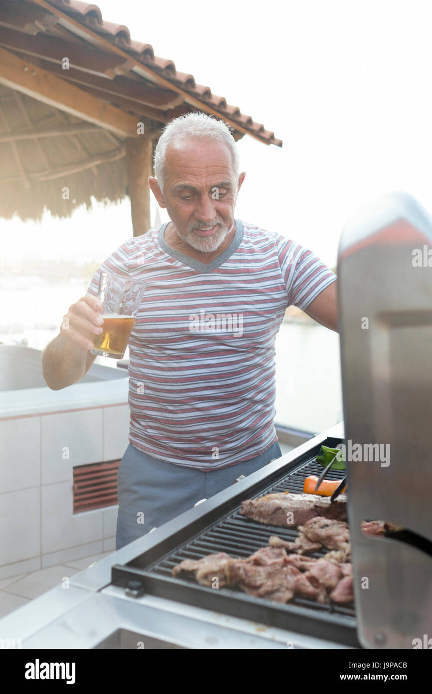Caucasian man autour de 60 griller la viande et les légumes sur un gril de gaz tout en appréciant un verre de bière Banque D'Images
