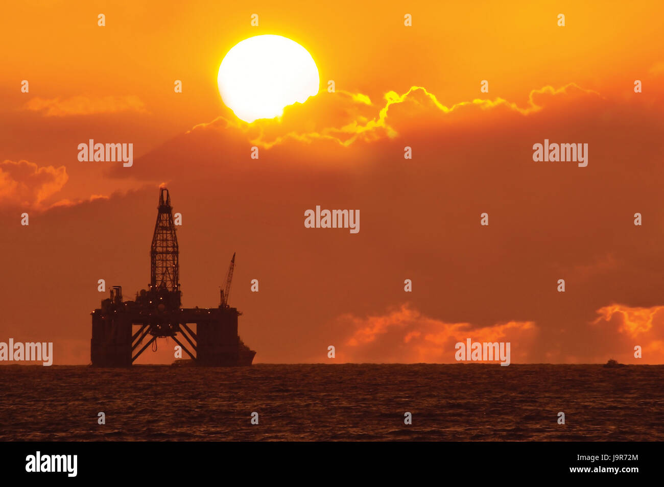 Coucher de soleil à travers les nuages, silhouetting une plate-forme pétrolière en mer. Banque D'Images
