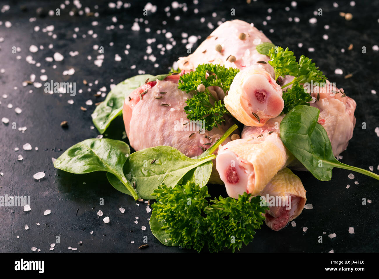 Photo horizontale de cuisses de poulet. Quelques morceaux de viande avec os. Les feuilles d'épinards et persil Vert sur noir vintage plateau avec sel, poivre, cumin un Banque D'Images