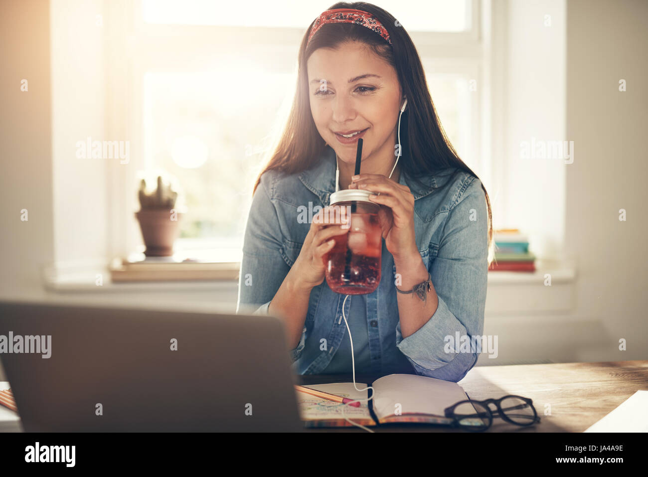 Young smiling businesswoman sitting avec boisson savoureuse et looking at laptop dans les écouteurs. Banque D'Images