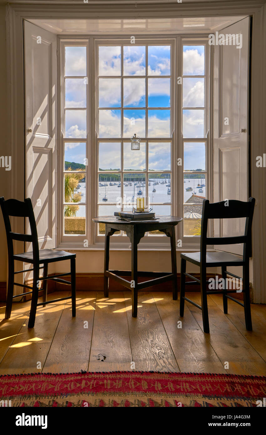 Deux chaises vides et une table ronde à l'intérieur d'un chalet à la  recherche à travers une grande fenêtre ensoleillée à une vue de bateaux  amarrés dans le port Photo Stock -