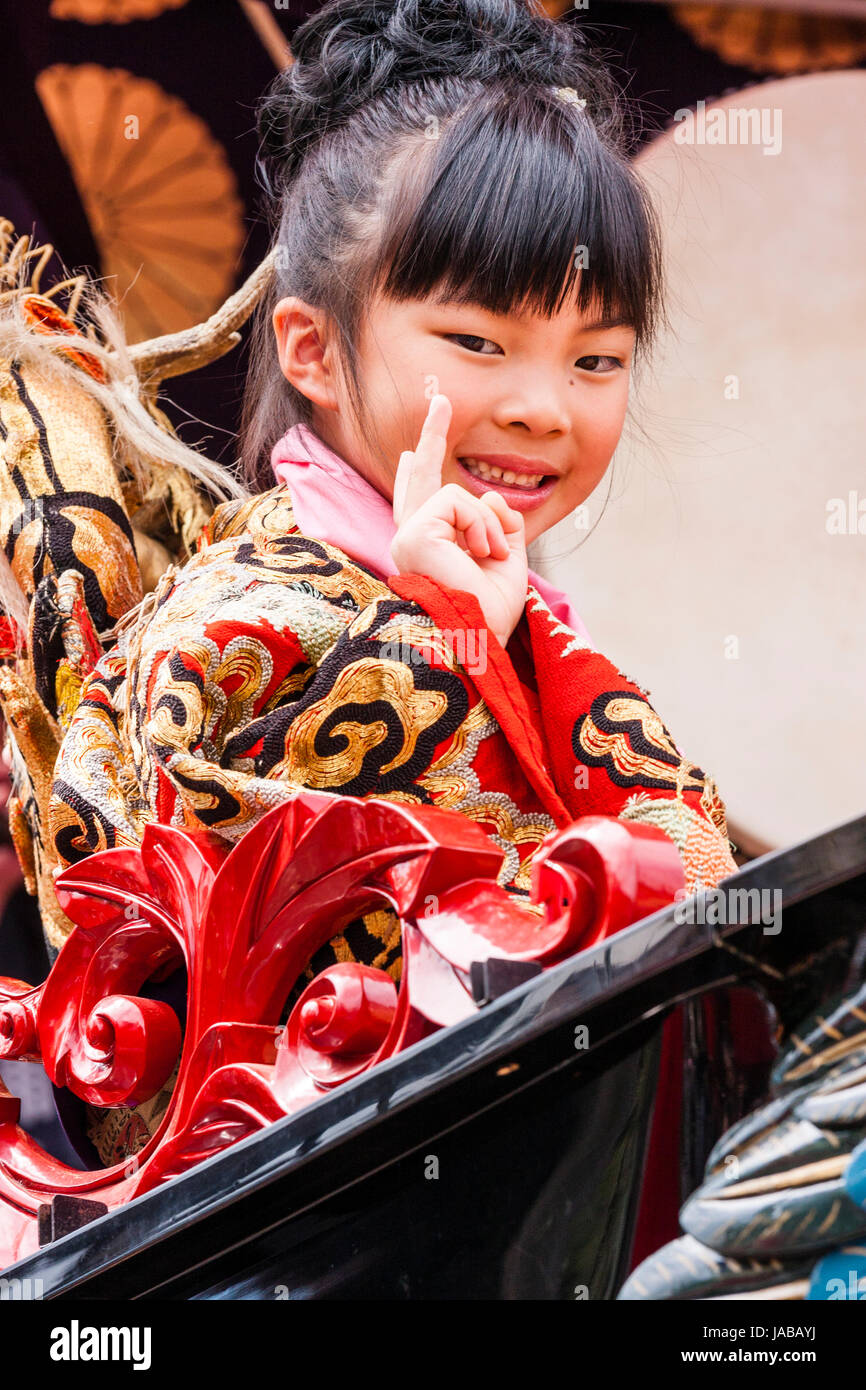 L'enfant japonais, fille, 8-9 ans, souriant et saluant avec deux doigt geste de paix tout en étant assis sur le dashi flotter. Porte kimono rouge et or. Banque D'Images