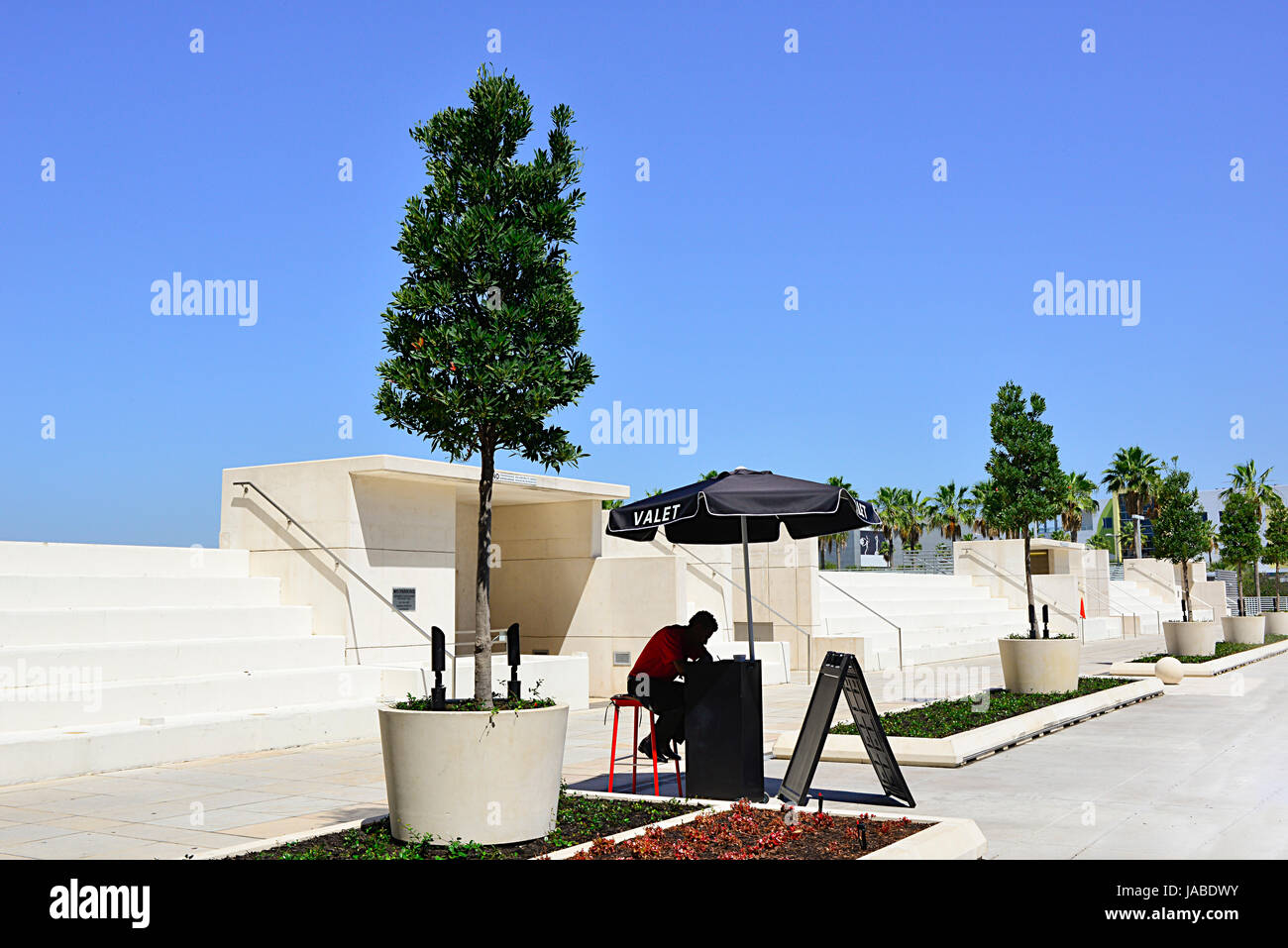 La silhouette d'un homme attendant valet assis sous un parasol dans un vaste terrain de stationnement au design contemporain blanc avec des arbres dans des jardinières Banque D'Images