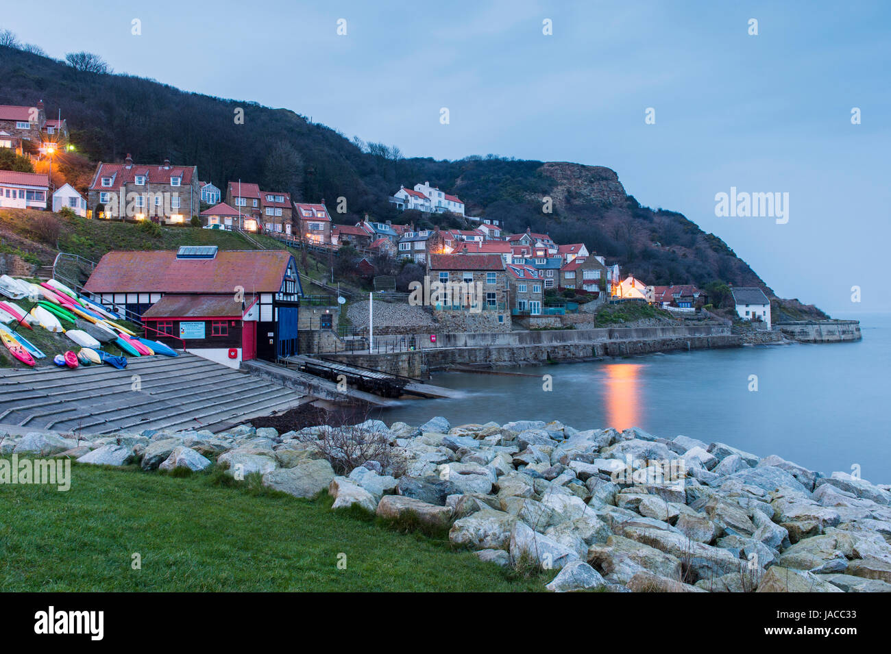 Les lumières sont allumées dans les maisons en vue de soirée joli village côtier pittoresque, avec des falaises, bateaux & mer calme - Runswick Bay, North Yorkshire, Angleterre, Royaume-Uni. Banque D'Images