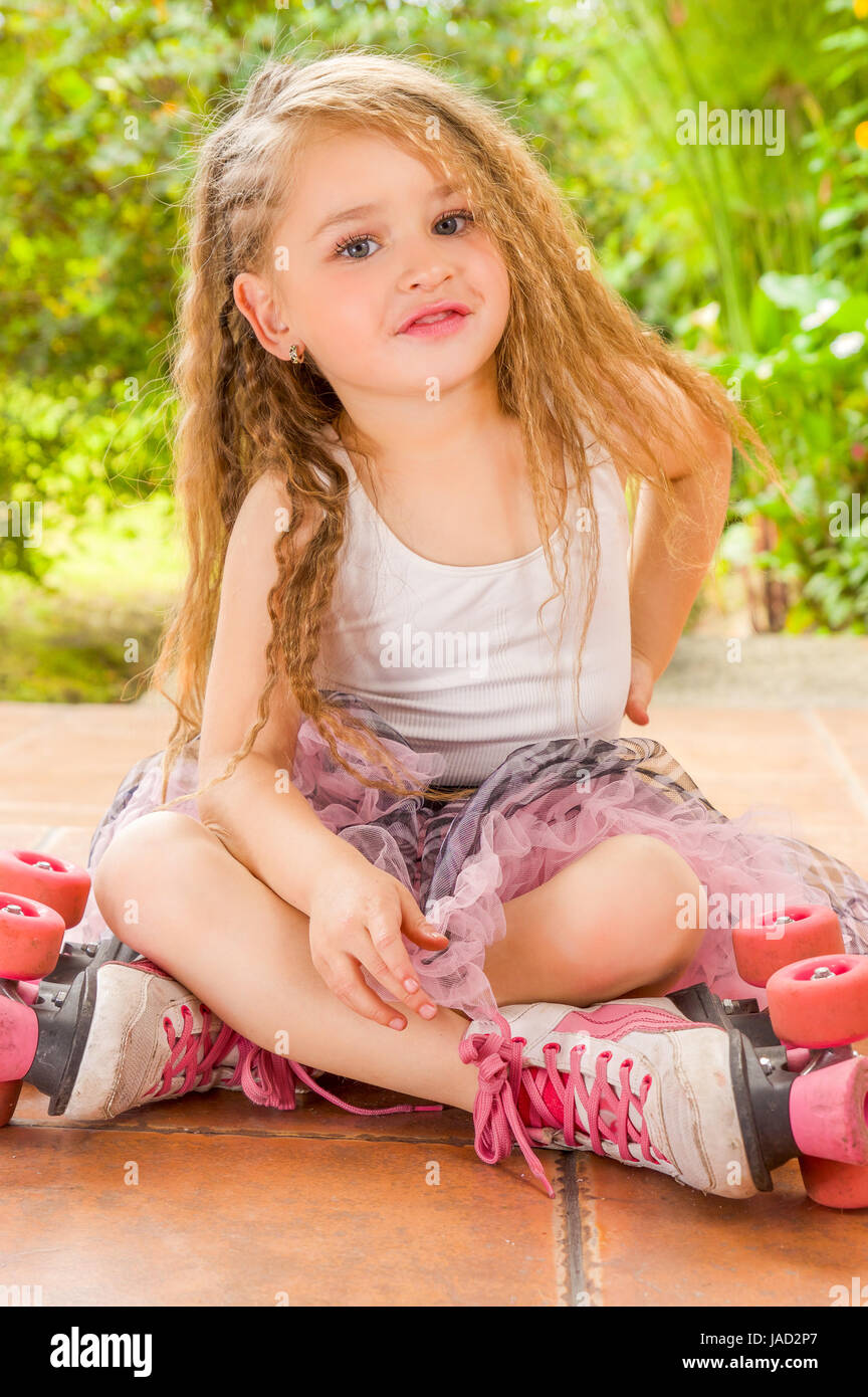 École maternelle Petite fille assise sur sol portant des patins à son passage et ses jambes, dans un fond de jardin Banque D'Images