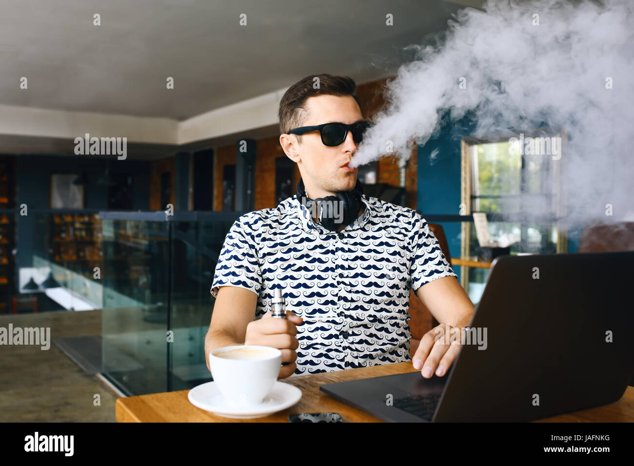 Beau jeune homme hipster avec barbe, assis dans un café avec une tasse de café, seine et libère un nuage d'vaporsitting. Au travail et d'avoir l'ordinateur portable Banque D'Images