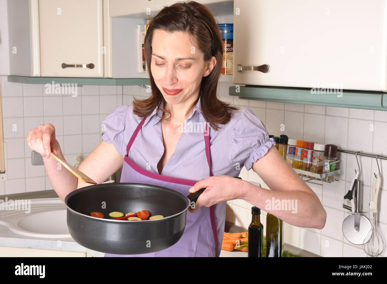 Erwachsene Hausfrau steht in ihrer hellen Einbauküche kocht frissen vágott kalódás gyertyán Gemüse in einer Pfanne. Banque D'Images