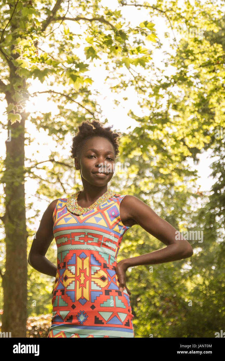 L'un, jeune adulte, black african american woman 29 ans, mains sur les hanches, portrait journée ensoleillée à l'extérieur, derrière les arbres, low angle shot, portant robe Banque D'Images