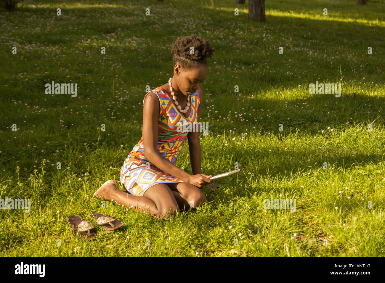L'un, jeune adulte, black african american woman 29 ans, assis, Tablette de lecture dehors parc herbe nature, journée ensoleillée à l'extérieur, elevated view, wea Banque D'Images