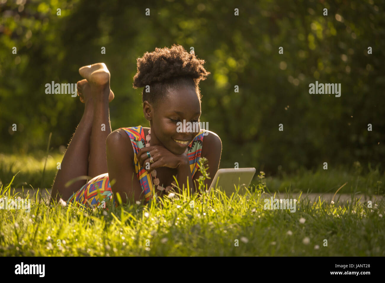 L'un, jeune adulte, black african american woman happy smiling bénéficiant à 29 ans, fixant lying on grass, Tablette de lecture dehors parc nature, sunny d Banque D'Images