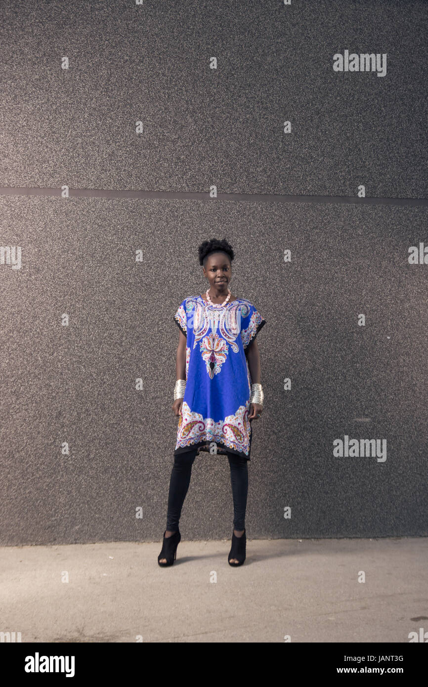 L'un, jeune adulte, black african american woman, 29 ans, vierge d'expression, à la caméra à l'extérieur, mur gris, portant robe bleue, collants, haut Banque D'Images