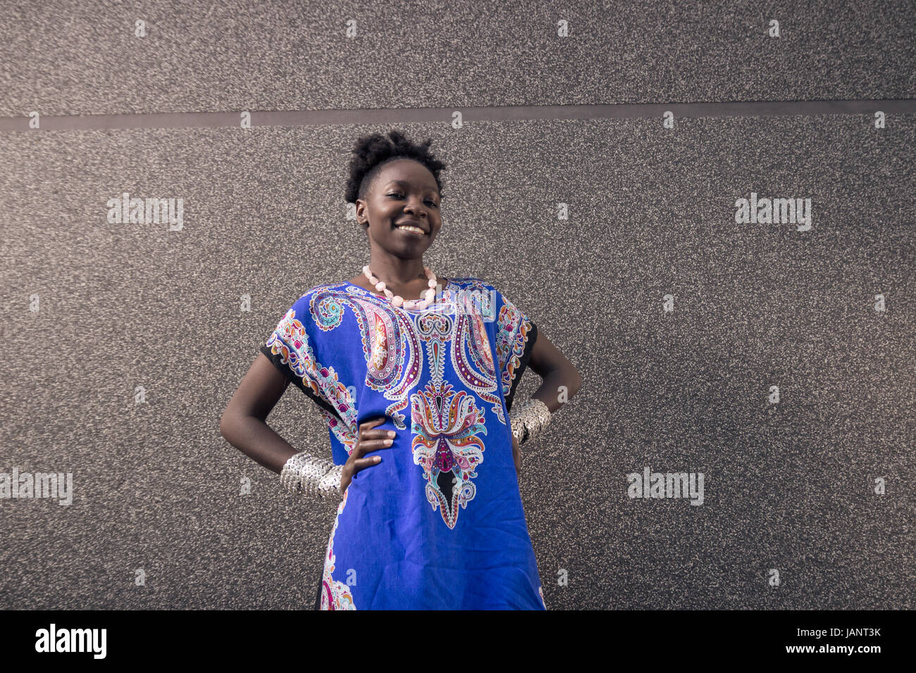 L'un, jeune adulte, black african american woman, 29 ans, à la caméra à l'extérieur, mur gris, portant robe bleue, smiling happy, mains sur les hanches Banque D'Images