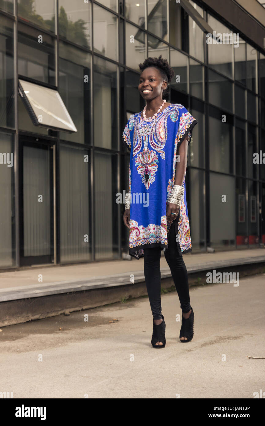 L'un, jeune adulte, black african american woman, 29 ans, souriant, chaussée de marche extérieur de l'immeuble, à la recherche d'appareil photo, à l'extérieur, wearing blue dr Banque D'Images