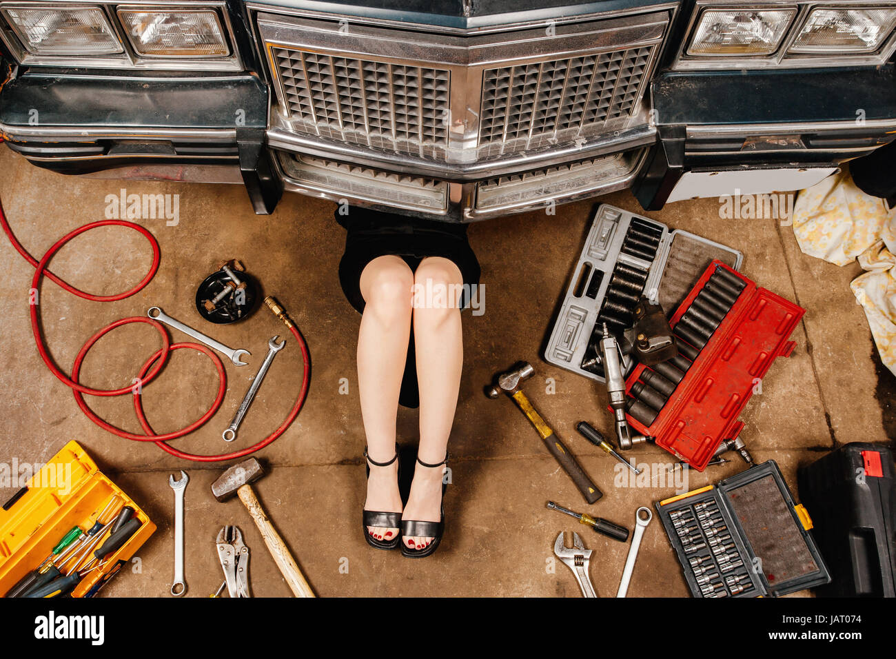 Une femme portant une jupe noire et des talons d'effectuer les réparations exigées en vertu de l'avant d'une vieille voiture du début des années 80. Banque D'Images