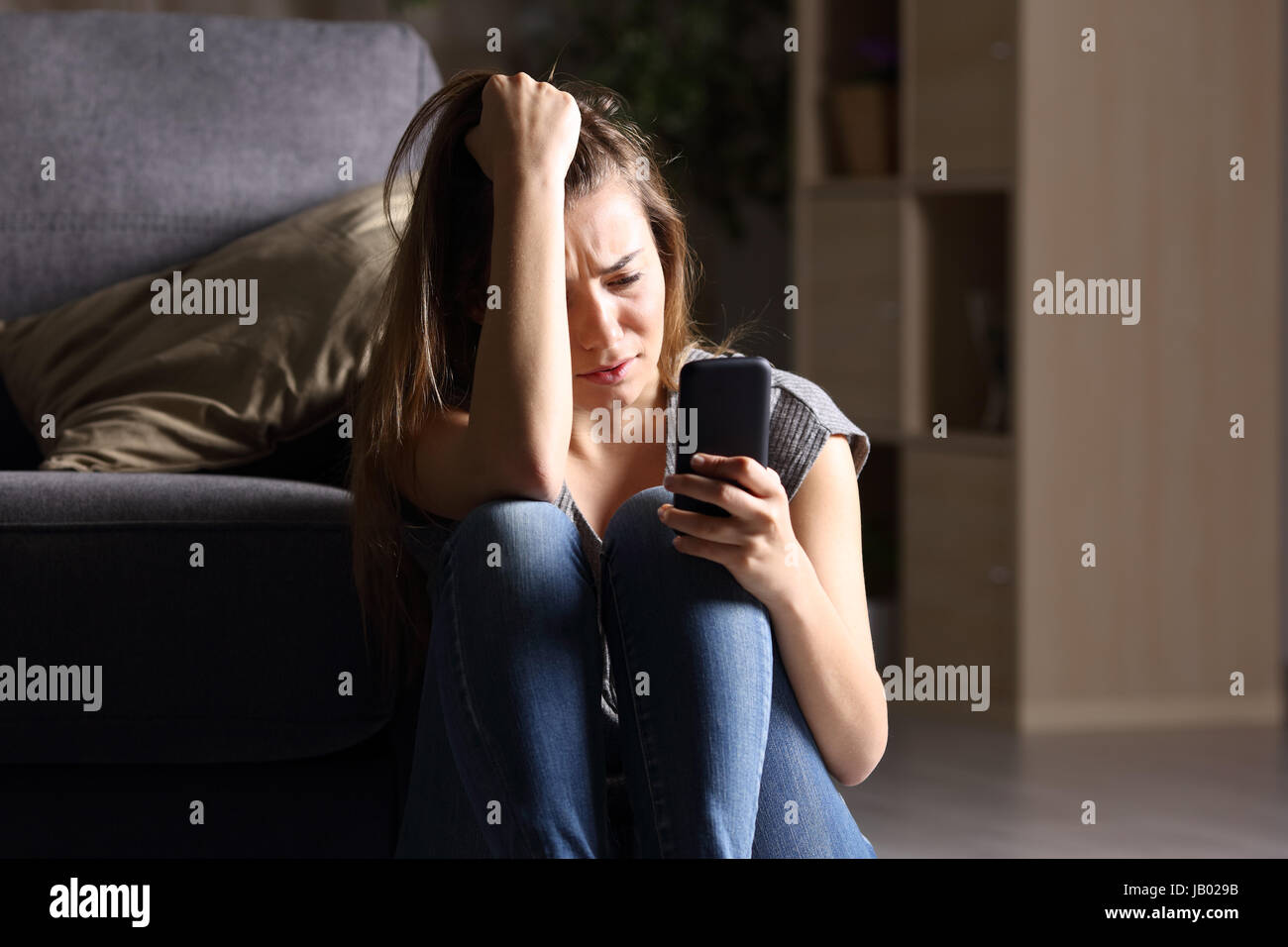 Vue frontale d'un triste teen phone assis sur le plancher dans la salle de séjour à la maison avec un fond sombre Banque D'Images