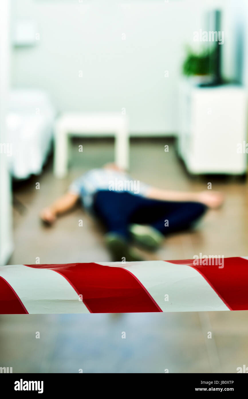 Détail d'une scène de crime avec le corps mort d'un jeune homme étendu dans le sol à l'intérieur, avec un ruban de police rouge et blanc à l'avant-plan Banque D'Images