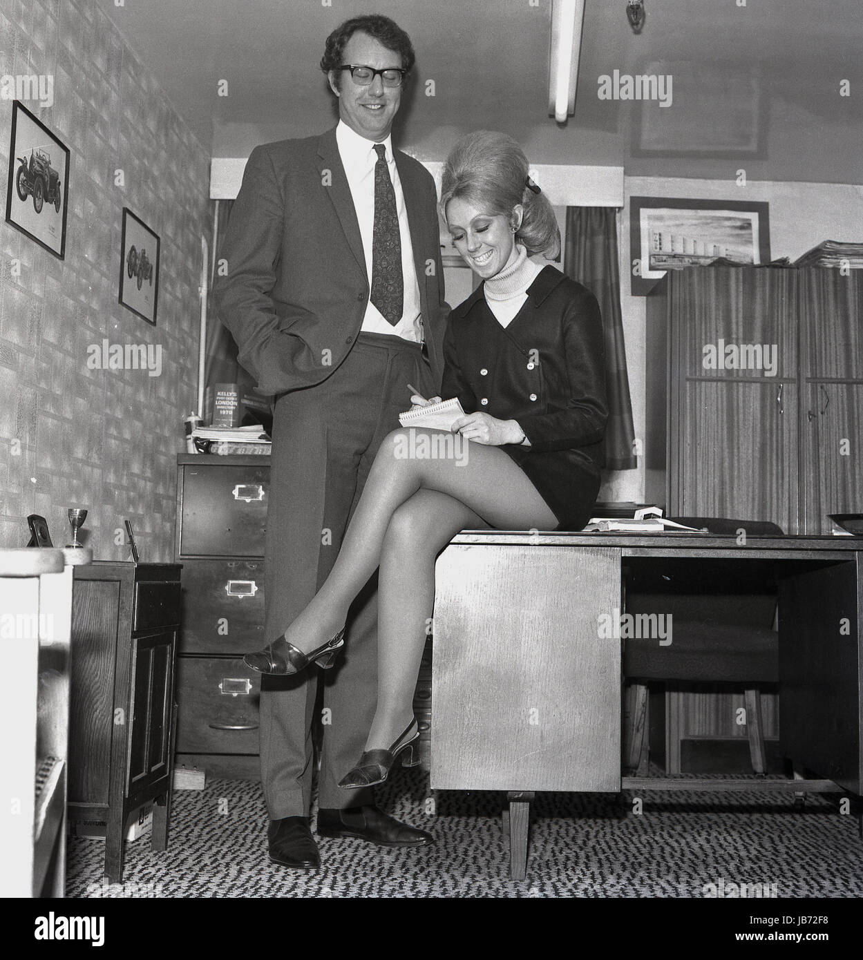 Années 1970, historique, d'un secrétaire portant un mini-shirt est assis  sur le bord d'un bureau alors qu'elle prend les notes vers le bas de son  patron dans son bureau Photo Stock -