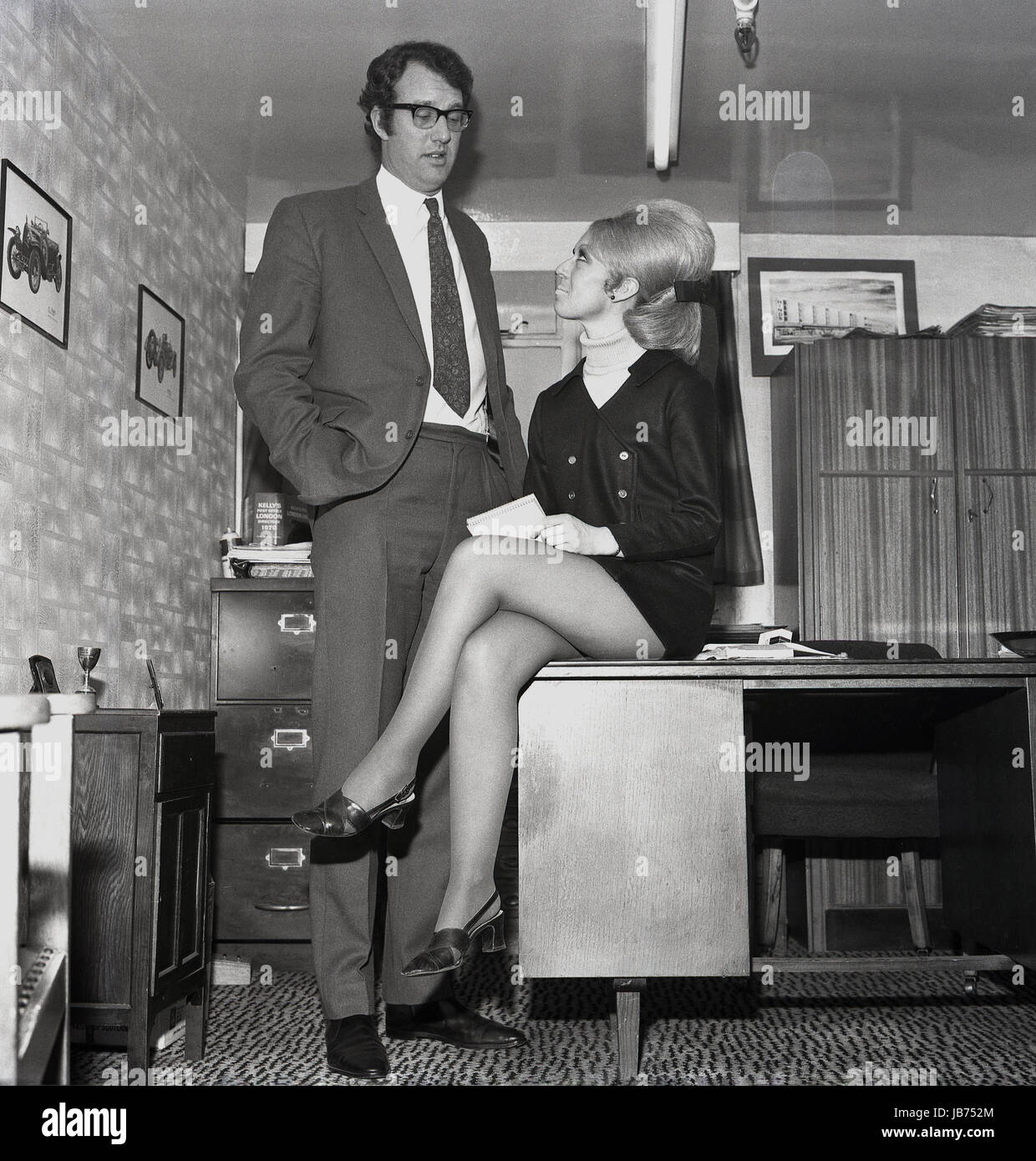 Années 1970, historique, d'un secrétaire portant une mini-jupe est assise  sur le bord d'un bureau alors qu'elle prend les notes vers le bas de son  patron dans son bureau Photo Stock -