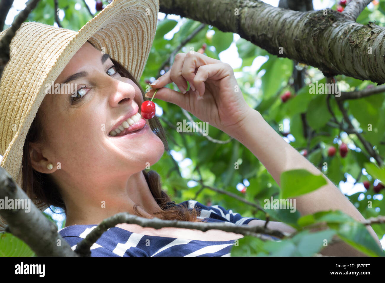 Une femme portant un chapeau sur un arbre de toucher une cerise avec sa langue. smiling, looking at camera. Banque D'Images