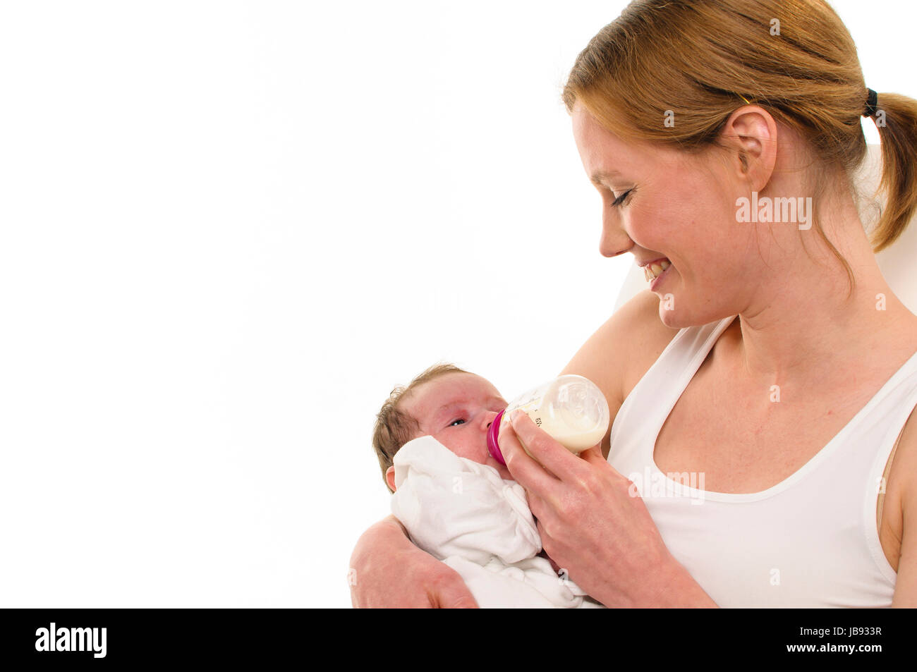 Erwachsene Eine Frau mit weißem Shirt hält einen gekleideten Säugling les weiß auf und zärtlich bras gibt ihm sein Fläschchen, freigestellt Hintergrund weißem vor. Banque D'Images