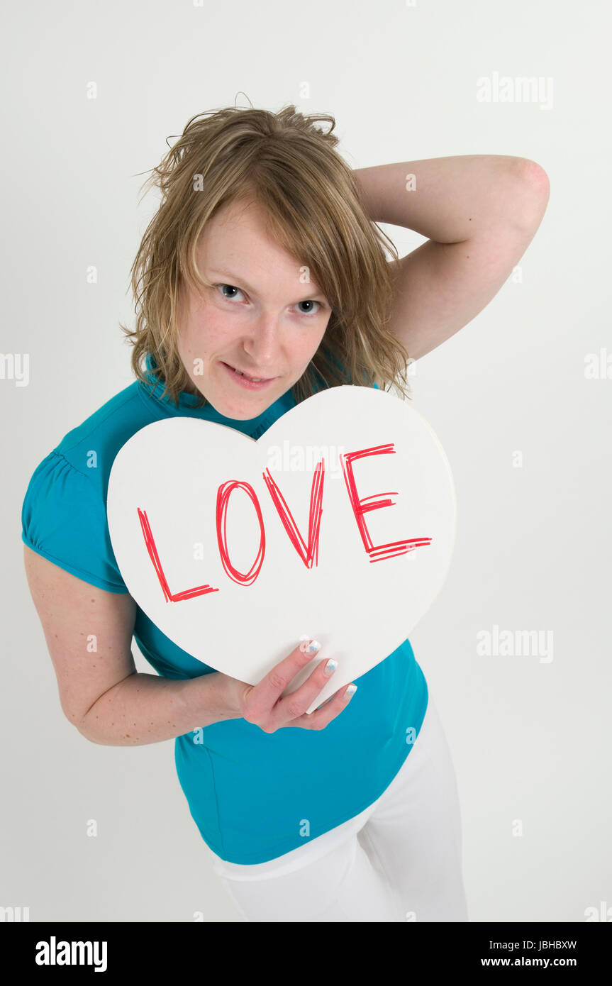 Oberkörperporträt einer jungen Frau im blondn hellblauen T-Shirt eine weisses Herz vor sich mit der Aufschrift haltend 'Amour' und mit der linken main dans das Eigene Haar greifend Banque D'Images